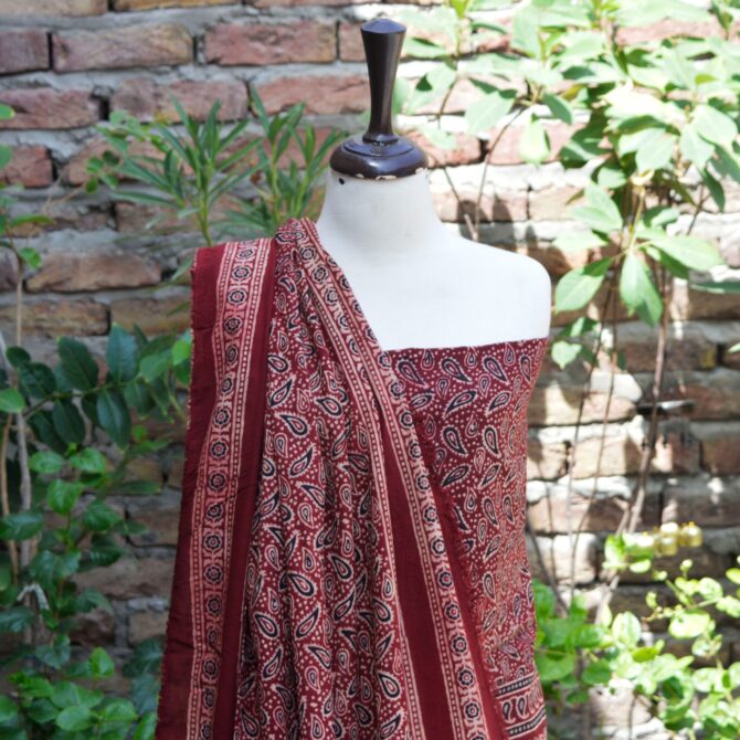 A folded maroonish red Ajrak un-stitched fabric, decorated with sharp, elegant hand block patterns, crafted with organic dyes, celebrating traditional artistry in a stylish, modern form.