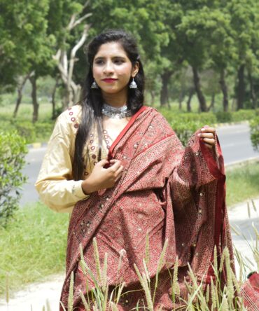 A model gracefully draped in the Organic Dye Hand Block Printed Ajrak Odhni Sassui, showcasing intricate geometric and floral patterns in Mehroonish Red, adorned with shimmering sitara sequences and thread tassels.