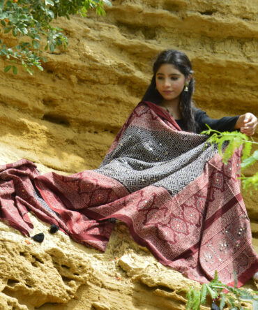 A model elegantly draped in the Organic Dye Hand Block Printed Ajrak Odhni Taskeen, showcasing intricate geometric and floral patterns in deep Black and Mehroonish Red, enhanced with shimmering sitara sequences and thread tassels.