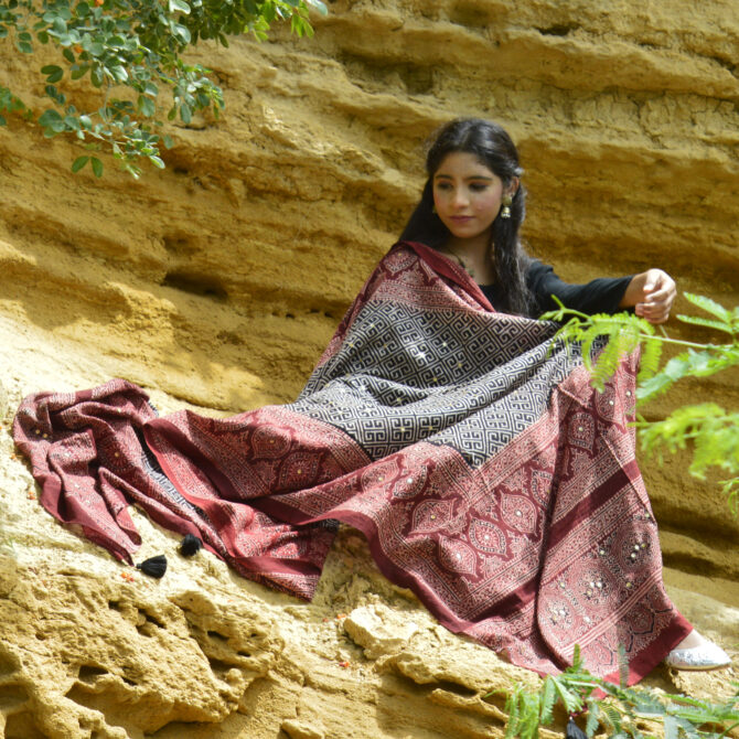 A model elegantly draped in the Organic Dye Hand Block Printed Ajrak Odhni Taskeen, showcasing intricate geometric and floral patterns in deep Black and Mehroonish Red, enhanced with shimmering sitara sequences and thread tassels.