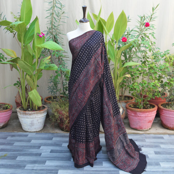 A beautifully handcrafted Ethnic Ajrak Saree with polka dot patterns in deep black, blending traditional Ajrak block printing with modern design. Made from breathable, eco-friendly Modal Silk with intricate geometric and floral motifs.